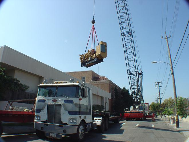 Caterpillar, 3512 diesel engine generator being craned into place across a large commercial building.